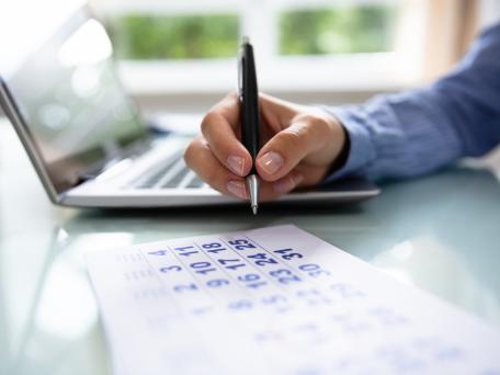 Person markiert Termin in Kalender neben geöffnetem Laptop.