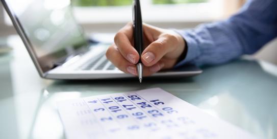 Person markiert Termin in Kalender neben geöffnetem Laptop.